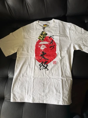 Vit BAPE t-shirt med japanskt tryck - Vit t-shirt från BAPE med ikoniskt rött ape-head tryck och japanska tecken på framsidan. Klassisk rund hals och korta ärmar. Märkets logga syns på etiketten och camo-taggen ger extra streetwear-känsla.