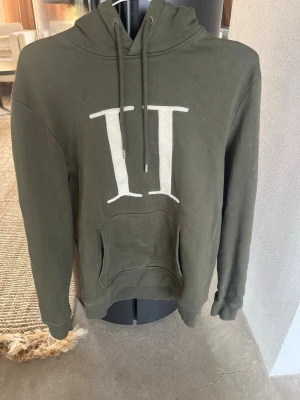 Olivgrön hoodie med vit logga - Snygg olivgrön hoodie med stor vit logga på bröstet. Klassisk känguruficka framtill och justerbar huva med dragsko. Perfekt för en avslappnad och trendig stil. Mjuk bomullskänsla och långa ärmar.