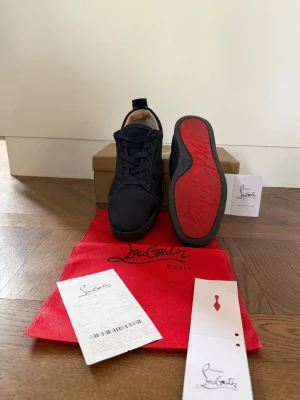 Sneakers Christian Louboutin Louis Junior Flat Suede Navy - Elegant Christian Louboutin Louis Junior Flat low-top sneakers in rich navy suede, featuring matching laces and a sleek black rubber sole. The iconic vibrant red outsole, a signature of the brand, is visible on the underside.  Condition : 6/10 (Visible wear and scuffs on red outsoles) Color : Navy Blue Fast and careful shipping 