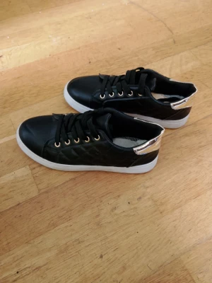 Svarta sneakers med guldiga detaljer - Snygga svarta sneakers med quiltad sida, vita sulor och guldiga metalldetaljer vid snörningen. Baktill finns en metallic guldig hälkappa som ger extra edge. Perfekta för dig som vill sticka ut lite extra i vardagen.