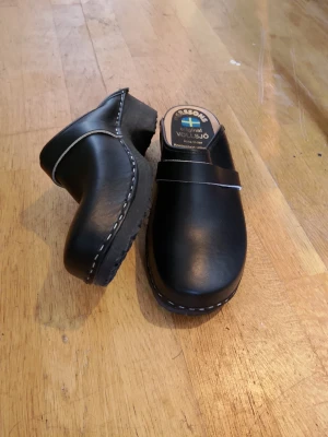 Svarta trätofflor från Larssons - Klassiska svarta trätofflor från Larssons Original Volsjö med ovandel i slätt läder och sula i trä. Snygg vit söm längs kanten och en enkel rem över foten. Perfekta för dig som gillar stilren och tidlös design.
