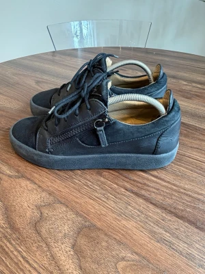 Svarta sneakers med dragkedja - Snygga svarta sneakers från Giuseppe Zanotti med lågt skaft, snörning och dragkedja på sidan. Skorna är gjorda i mocka och läder med en robust gummisula. Perfekta för dig som gillar stilrena och trendiga skor med en edge.