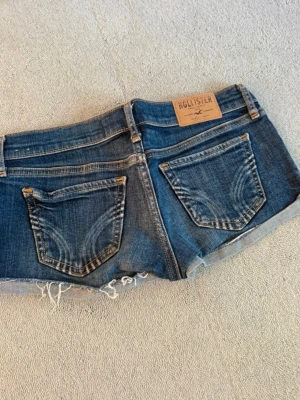 Blå jeansshorts från Hollister - Snygga blå jeansshorts från Hollister med råa, fransiga kanter och klassiska fem fickor. Shortsen har låg midja och är tillverkade i bomull. Perfekta för sommardagar och en avslappnad stil.