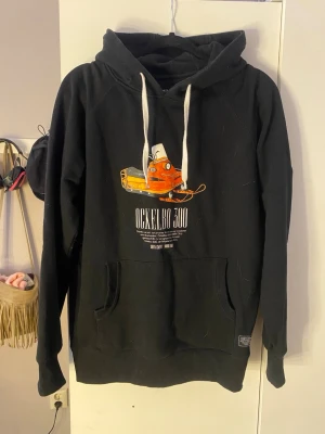 Svart hoodie Ockelbo 300 Sortn Company - Svart hoodie med tryck av en orange snöskoter och texten 'Ockelbo 300' på bröstet. Stora vita dragsnören i huvan och magficka fram.