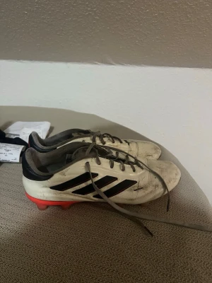 fotbollsskor  - Säljer ett par vita fotbollsskor från Adidas med svarta ränder och röda dobbar. Skorna har klassisk snörning och är tillverkade i syntetmaterial. Perfekta för spel på gräsplan och har en sportig look som sticker ut på planen.