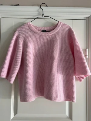 Ljusrosa stickad tröja med halvärm - Superfin ljusrosa stickad tröja med halvärmar och rund halsringning. Tröjan har en mjuk och fluffig känsla och är ribbstickad för extra struktur. Perfekt för lager-på-lager och en söt touch till din outfit.