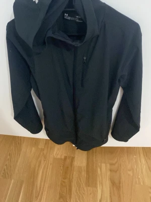 Svart vindjacka från Under Armour - Svart vindjacka från Under Armour med huva och dragkedja framtill. Jackan är i sportig modell och tillverkad i syntetmaterial, perfekt för kyligare dagar. Diskret logga på bröstet och smidig passform.
