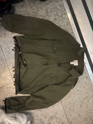 Olivgrön jeansjacka med bollar - Snygg olivgrön jeansjacka med två bröstfickor och dekorativa färgglada bollar längs nederkant och ärmslut. Klassisk krage och knäppning framtill. Perfekt för dig som vill sticka ut med en unik detalj på jackan.