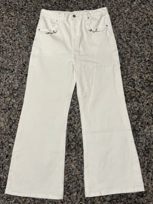 Vida vita jeans med detaljer - Säljer ett par vita jeans med vida ben och snygga silverdetaljer vid fickorna fram. Jeansen har två bakfickor och ett justerbart spänne bak i midjan. Tillverkade i ett kraftigare bomullstyg som ger en clean och fräsch look.