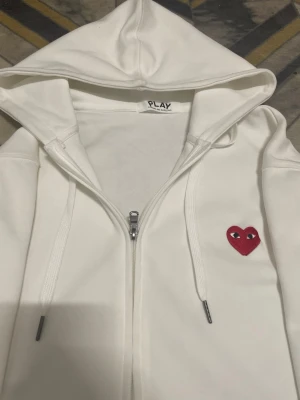 Vit hoodie från Comme des Garçons Play - Vit hoodie med dragkedja från Comme des Garçons Play. Har huva med dragsko och ett rött hjärta med ögon broderat på bröstet. Mjuk bomullskänsla och klassisk passform, perfekt för en clean och stilren look.