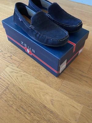 Marinblå loafers från Zeus - Stilrena marinblå loafers från Zeus i mocka med vita kontrastsömmar och randigt innerfoder. Nästan helt nya, har använt dom 2 gånger!! Box medföljer. Storleken är 43 men skulle säga att dom passar 44! Perfekt till sommaren. Pris kan diskuteras!