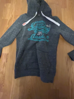 Mörkgrå hoodie från Superdry - Mörkgrå hoodie från Superdry med turkos textprint på bröstet och vita dragsnören i huvan. Klassisk känguruficka framtill och mjukt, fleeceliknande innerfoder. Perfekt för en avslappnad streetstil.skriv privat för frågor priset är ej hugget i sten 