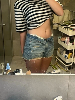 Blå jeansshorts med slitna detaljer - Säljer ett par blå jeansshorts med slitna detaljer och fransig kant. Perfekt att vika ner och ha över bikinitrosor på stranden. Korta och höga i midjan annars så passar också med en stickad tröja över på sommarkvällar. 
