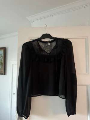 Svart blus med spets och volang H&M - Svart blus från H&M, spetsdetaljer vid halsringningen och volang längs bröstet. Blusen är luftig och väldigt skön att ha