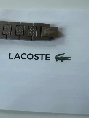 Silverfärgat armband från Lacoste - Stilrent armband i silverfärgad metall från Lacoste. Länkarna har en mix av släta och texturerade rutor, och spännet pryds av den klassiska krokodilloggan. Perfekt accessoar för dig som gillar en clean och modern look.