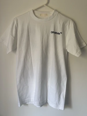Off-White vit t-shirt med grafiskt tryck - Vit t-shirt från Off-White i bomull med svart logga på bröstet och stort grafiskt tryck på ryggen med tegelmönster och figur. Klassisk rund hals och korta ärmar, perfekt för en streetwear-look.