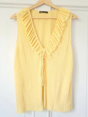 Gul plisserad blus med volang från Zara - Superfin gul plisserad blus från Zara med djup v-ringning, volangkant och knyt framtill. Ärmlös modell i lätt och luftig polyester, perfekt för varma dagar. Endast använd 1 gång! Såå fin till sommaren!