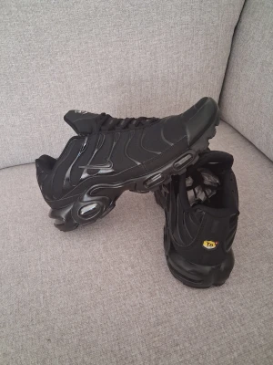 Nike Air Max Plus Triple Black - Säljer ett par Nike Air Max Plus i färgen Triple Black. Skorna har en sportig design med mesh och syntetmaterial, ikoniska Tn-detaljer på hälen och synlig Air-dämpning i sulan. Perfekta för dig som gillar streetwear och vill ha en clean look.