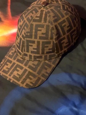 Fendi keps med FF-mönster - Snygg keps från Fendi med det klassiska bruna och svarta FF-logomönstret över hela kepsen. Justerbar rem baktill i mörkbrunt läder med Fendi-logga. Perfekt accessoar för att lyfta din streetwear-look.