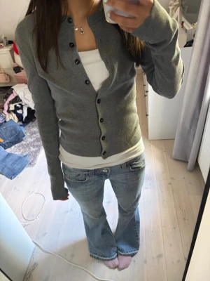 Grå kofta - En stilren grå kofta med långa ärmar och svarta knappar framtill. Modellen är figurnära och passar perfekt över ett linne eller t-shirt. Koftan har en klassisk rund halsringning och är tillverkad i ett mjukt, tunt stickat material. Knappt använd så som ny. Tveka inte att fråga ifall du undrar något!