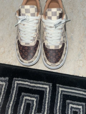 Louis Vuitton-inspirerade sneakers - Snygga sneakers med klassiskt Louis Vuitton-mönster i brunt och beige. Skorna har rutigt mönster på ovandelen och bruna partier med LV-logga. Vita snören och beige insida. Perfekta för dig som vill sticka ut med lyxig streetstyle.
