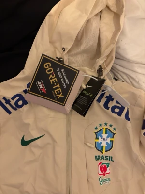 Nike Brasil vindjacka GORE-TEX beige - Snygg beige vindjacka från Nike med Brasil-logga och flera sponsortryck. Jackan har huva, hel dragkedja och är tillverkad i GORE-TEX-material som andas. Perfekt för dig som gillar sportig stil och vill ha något unikt från landslaget.