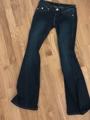 True Religion mörkblå bootcut jeans st w24 - Mörkblå bootcut jeans från True Religion med klassiska kontrastsömmar och snygga detaljer på bakfickorna. Jeansen har fem fickor, orangea sömmar och en ikonisk logga på bakfickan. Perfekta för dig som gillar en tidlös och cool stil.