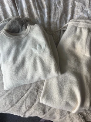 Krämvitt loungewear-set från Adidas - Mysigt loungewear-set från Adidas i krämvit färg. Setet består av en långärmad tröja med ribbade muddar och matchande byxor med resår i midjan. Använda under vintern. 