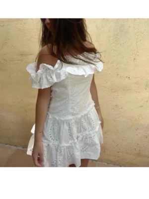 Vit offshoulder klänning med volang - Superfin vit kortklänning med offshoulder och volangdetaljer. Klänningen har broderade mönster och är tillverkad i bomull, vilket gör den luftig och perfekt för varma dagar. Den har en romantisk och somrig vibe med sin omlottliknande kjol och rynkade detaljer.