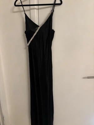 Svart långklänning med strassdetalj Zara  - Elegant svart långklänning i satinliknande material med smala axelband och en snedställd strassdetalj framtill. Klänningen har en figurnära passform och djup v-ringning, perfekt för dig som vill sticka ut på festen.