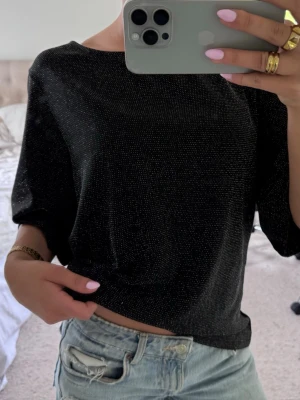 Svart glittrig topp med halvärm - Svart topp med glittriga detaljer och halvärm. Modellen är lite loose och toppen har en rund halsringning. Perfekt för dig som gillar en enkel men ändå festlig stil.