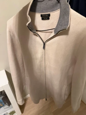 Beige ziptröja från Massimo Dutti - Stilren beige ziptröja från Massimo Dutti med hög ribbad krage i grått. Tröjan har långa ärmar, två fickor framtill och är tillverkad i ett mjukt stickat material. Perfekt för lager-på-lager och enkel att matcha med olika outfits.