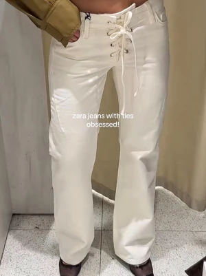Vita jeans med snörning från Zara - Säljer ett par vita jeans från Zara med unik snörning framtill istället för vanlig knapp och dragkedja. Jeansen har raka ben och låg midja, vilket ger en snygg och trendig siluett. Perfekta för dig som vill sticka ut med detaljer! Materialet är denim.
