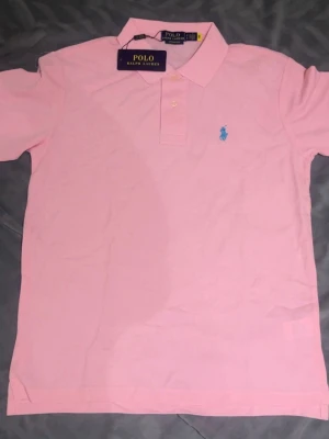 Ljusrosa pikétröja från Ralph Lauren - Snygg ljusrosa pikétröja från Polo Ralph Lauren med klassisk krage och två knappar framtill. Tröjan har korta ärmar och den ikoniska blå broderade loggan på bröstet. Perfekt för en stilren och avslappnad look.