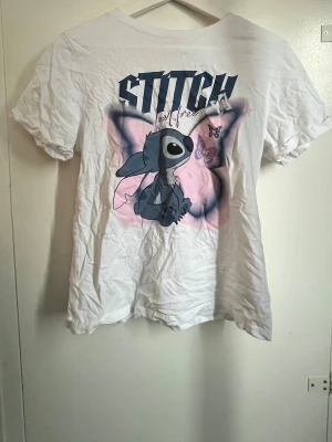 Vit Stitch t-shirt med stitch tryck - Säljer en vit t-shirt med stort tryck av Stitch från Disney framtill. T-shirten har korta ärmar och en avslappnad passform. Perfekt för dig som älskar tecknade motiv och vill ha en unik look. Också perfekt till ett barn :) 