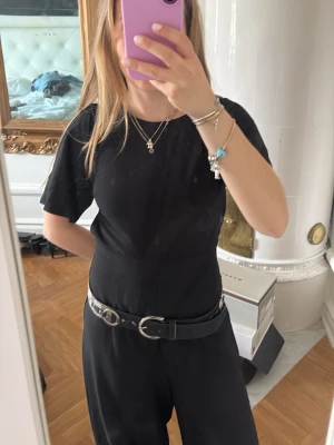 Svart byxdress - En svart byxdress med korta ärmar, rund hals och vida ben. Midjan markeras snyggt med formen och crossovern baktill, går även att bara knyta. Byxdressen har en clean och stilren look som är lätt att styla med smycken och accessoarer.