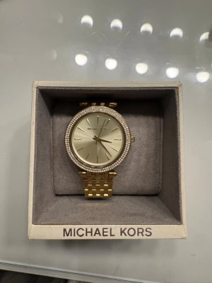Guldig klocka från Michael Kors - Stilren guldig klocka från Michael Kors med länkarmband och glittrande stenar runt urtavlan. Använd fåtals gånger. Man får även med extra knipps så man kan göra den större och mindre. 