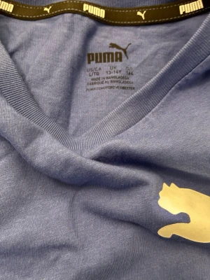 Blå Puma t-shirt med logga - Snygg blå t-shirt från Puma med klassisk svart och vit logga på bröstet. T-shirten har rund halsringning och är kortärmad, perfekt för en avslappnad och sportig stil. Tillverkad i mjuk bomull som känns skön mot huden.