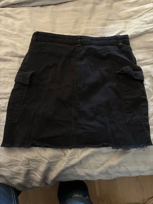 Svart kortkjol med fickor - Svart kortkjol i bomull med rå kant nertill och stora fickor på sidorna. Kjolen har knapp och dragkedja framtill och en enkel, rak passform. Perfekt för en avslappnad och cool stil.