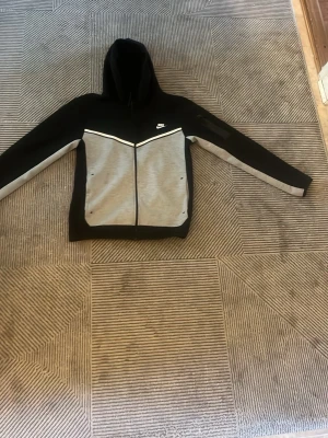 Nike hoodie svart och grå med dragkedja - Snygg hoodie från Nike i svart och grått med hel dragkedja framtill. Tröjan har huva, vita detaljer och två stora fickor på magen. Perfekt för en sportig och avslappnad stil. Nike-logga på bröstet och kontrastpaneler för extra edge.