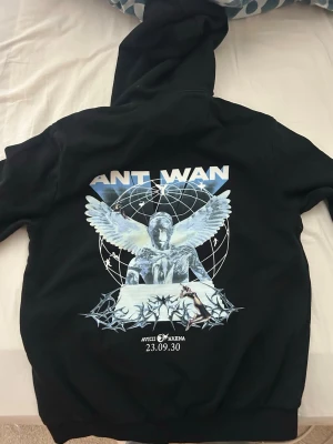 Svart Ant Wan hoodie med tryck - Svart hoodie med stor grafisk print på ryggen föreställande en ängel, taggtråd och texten 'Ant Wan' samt 'Avicii Arena 23.09.30'. Framsidan har en mindre logga på bröstet. Klassisk känguruficka och dragsko i huvan. Perfekt för dig som gillar streetwear och unika motiv.