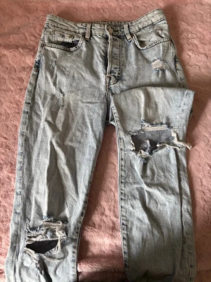 Ljusa ripped mom jeans i denim - Säljer ett par ljusblå mom jeans med ultra high waist och stora slitningar på benen. Jeansen är i klassisk denim och har en avslappnad passform med fem fickor och dragkedja. Perfekta för en trendig och avslappnad look.