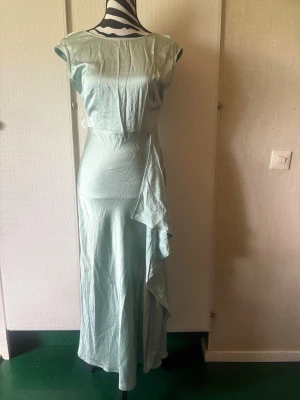 Ljusblå långklänning med volang - Elegant ljusblå långklänning i glansigt material med vacker drapering och volangdetalj framtill. Klänningen har korta ärmar och en djup v-ringning i ryggen som ger en snygg siluett. Perfekt för speciella tillfällen.