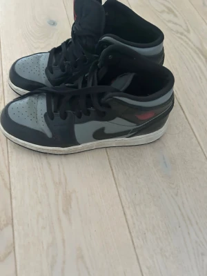 Nike Air Jordan 1 Mid svart/grå - Snygga Nike Air Jordan 1 Mid sneakers i svart och grått med klassisk svart swoosh och röd logga på sidan. 