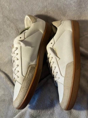 Vita Saint Laurent sneakers med gummisula - Snygga vita sneakers från Saint Laurent med klassisk låg profil och gummisula i brunt. Ovandelen är i perforerat läder och detaljer i beige mocka. Diskret Saint Laurent-logga på sidan och tungan. Perfekt för dig som vill ha stilrena och exklusiva skor. Modell SL10, nypris ca 8000kr. Storlek 43 passar 43,5 har blivit lagade för fram på den högra skon på mockan