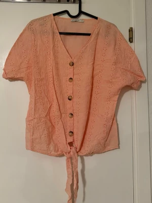 Persikofärgad blus från LCW Casual - Söt persikofärgad blus från LCW Casual med broderade blommor, v-ringning och knappar framtill. Blusen har korta ärmar och knytning nedtill för en trendig look. Perfekt för vår och sommar!