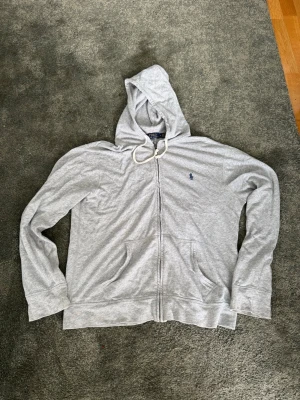 Grå hoodie från Polo Ralph Lauren - Snygg grå zip-hoodie från Polo Ralph Lauren med klassisk huva och dragsko. Broderad logga i blått på bröstet och två stora fickor framtill. Tillverkad i mjuk bomullsblandning för extra komfort. Perfekt för en chill och avslappnad stil.