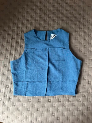 Blå ärmlös linnetopp  - Snygg blå linnetopp med hög ärmlös design. Toppen har en rak passform och stängs med knapp i nacken. Perfekt att styla med jeans eller kjol för en fräsch look.
