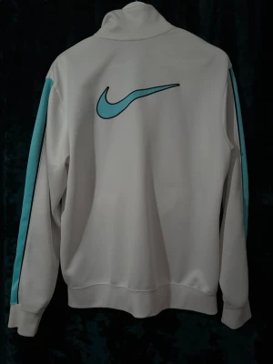 Vintage Vit Nike ziptröja med blå detaljer och stort Nike märke på ryggen - Snygg vintage vit ziptröja från Nike med ljusblå ränder längs ärmarna och klassisk Nike-logga på bröstet. Tröjan har hög krage, dragkedja framtill och två fickor med dragkedja. Perfekt för träning eller chill. Tillverkad i mjukt och bekvämt material.