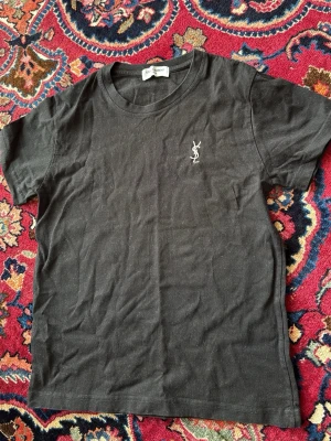 Svart t-shirt från Saint Laurent - Svart t-shirt från Saint Laurent med klassisk rund halsringning och vit YSL-logga broderad på bröstet. Enkel och stilren design som passar perfekt till jeans eller kjol. Tillverkad i mjuk bomull för en skön känsla hela dagen.